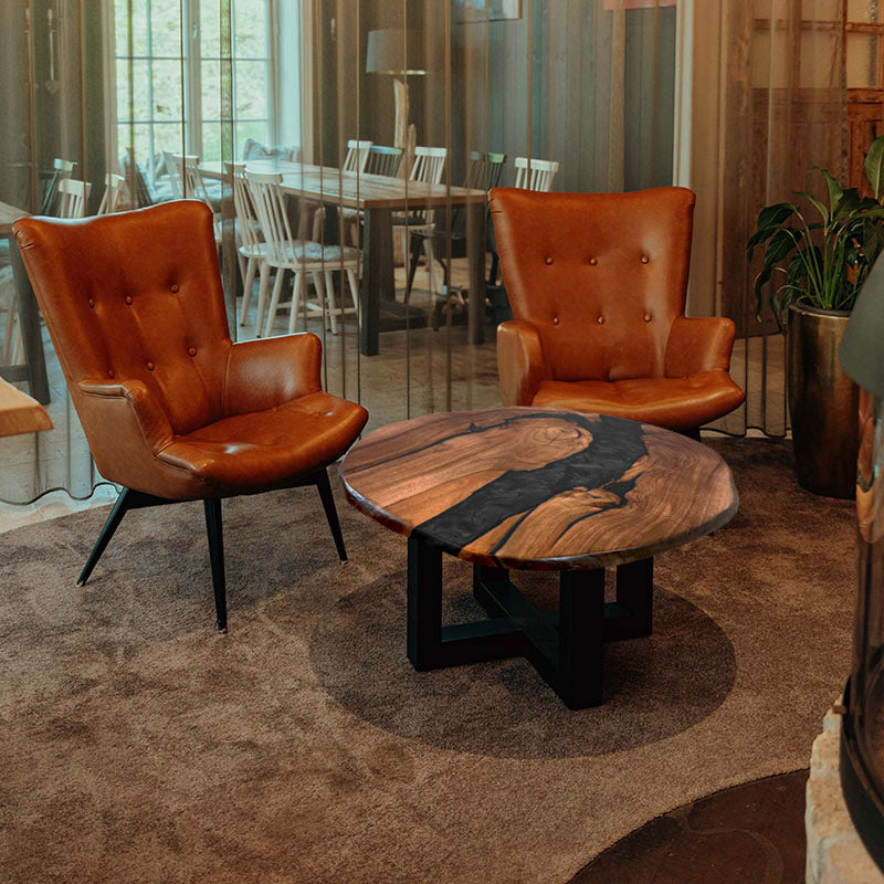 Couchtisch rund mit Naturkanten aus Nussbaum & Epoxidharz in blau, in gemütlichem Setting mit zwei braunen Ledersesseln. Modell Titisee von schwarzhirsch