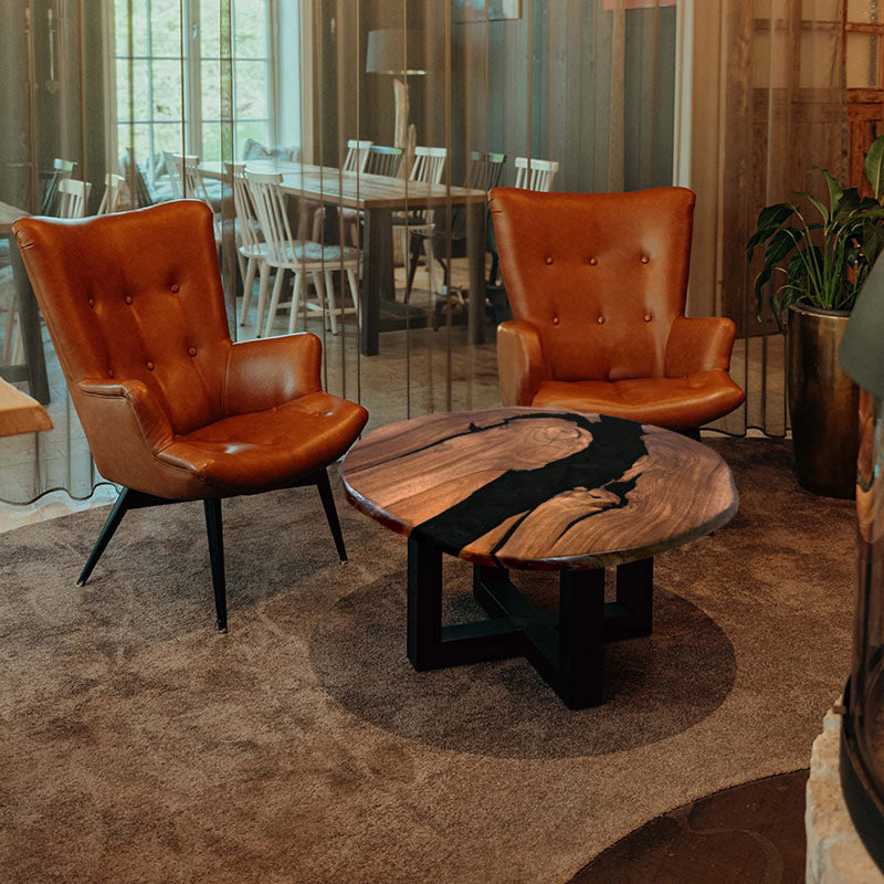 Couchtisch rund mit Naturkanten aus Nussbaum & Epoxidharz in schwarz, in gemütlichem Setting mit zwei braunen Ledersesseln. Modell Titisee von schwarzhirsch