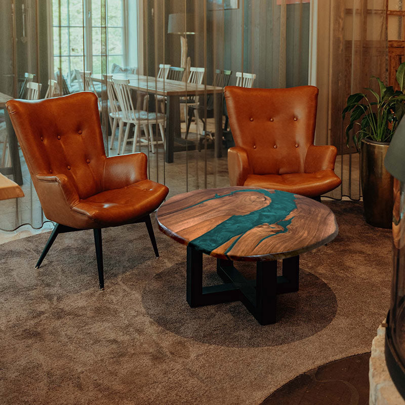 Couchtisch rund mit Naturkanten aus Nussbaum & Epoxidharz in türkis, in gemütlichem Setting mit zwei braunen Ledersesseln. Modell Titisee von schwarzhirsch