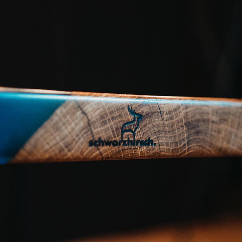 Tisch Epoxidharz. Detailbild Brennstempel mit schwarzhirsch Logo. Epoxidharz Tisch aus rustikaler Eiche mit blauem Epoxidharz. Modell Adria von schwarzhirsch