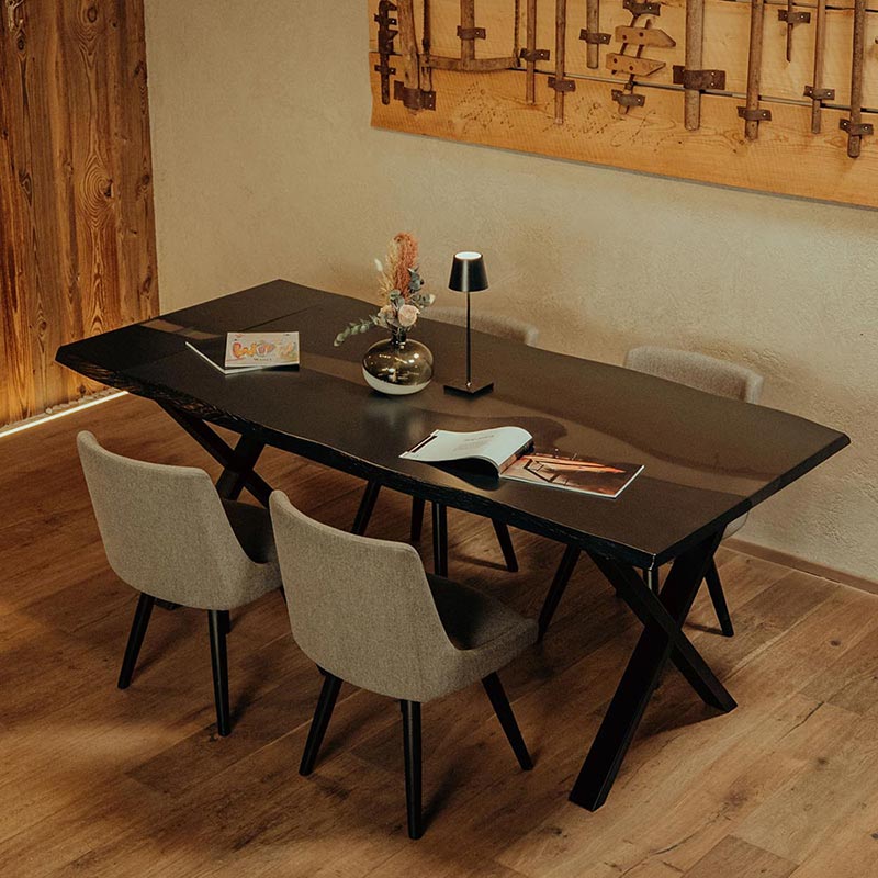 Esstisch aus schwarzer Eiche mit weißem Epoxidharz und Kreuzgestell. Der Tisch ist dekoriert mit einer Vase mit Trockenblumen und einer schwarzen Tischlampe. 4 graue Stühle umgeben den Tisch. Modell Blackforest von Schwarzhirsch.
