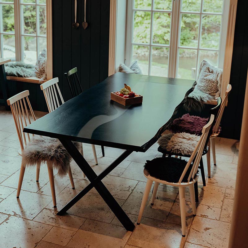 Esstisch schwarze Eiche mit weißem Epoxidharz und Kreuzgestell. Der Tisch ist dekoriert und gesäumt von 6 Stühlen. Modell Blackforest von Schwarzhirsch.